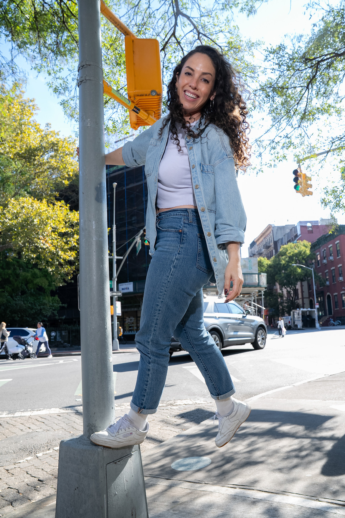 Woman in denim outfit smiles while hanging from a street pole on a sunny day in an urban city intersection with cars, buildings, trees, and pedestrians in the background.