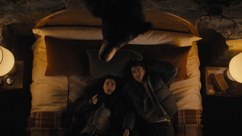 Two people lie side by side on a bed in a cozy, dimly lit room with rustic decor, lamps on nightstands, and a bear head mounted on the wall above them, suggesting a cabin or lodge setting.