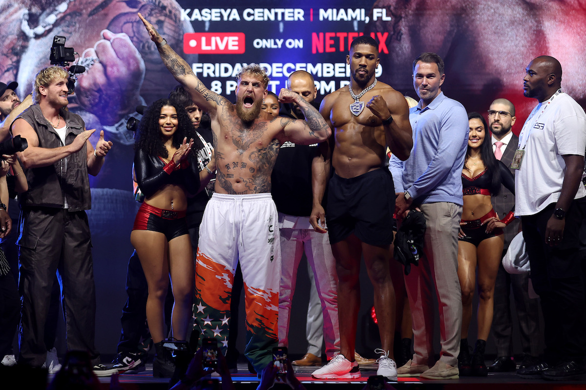 Jake Paul and Anthony Joshua pose shirtless on stage during a weigh-in event, surrounded by their teams, security, ring girls, and media, under bright lights with a promotional banner in the background displaying event details.