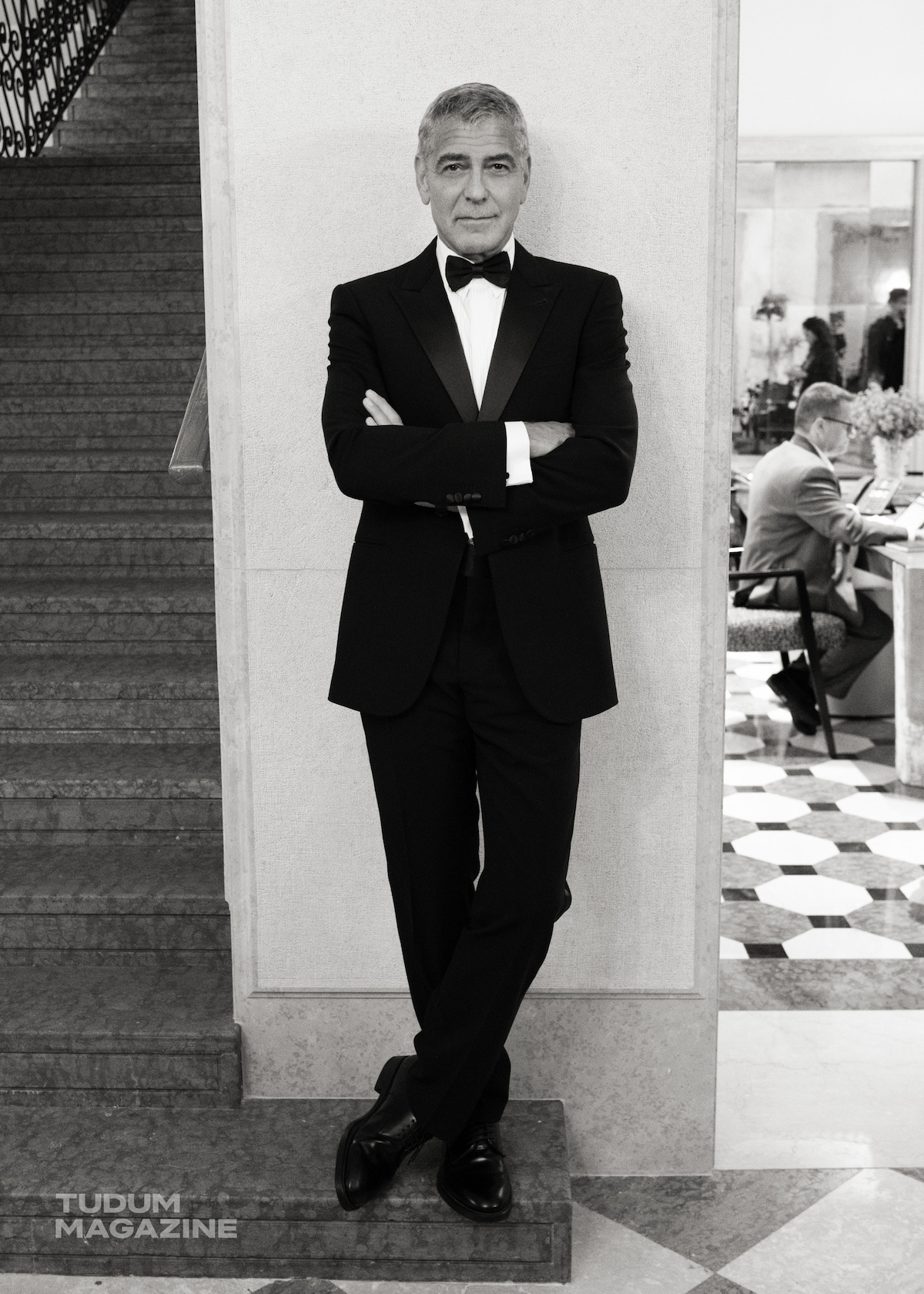 George Clooney stands at the foot of a staircase in a tuxedo.