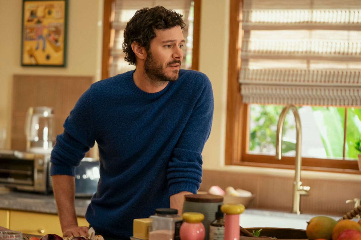 A man with curly hair and a beard wearing a blue sweater stands in a cozy kitchen leaning on the counter, surrounded by cooking ingredients, with sunlight coming through the window blinds.