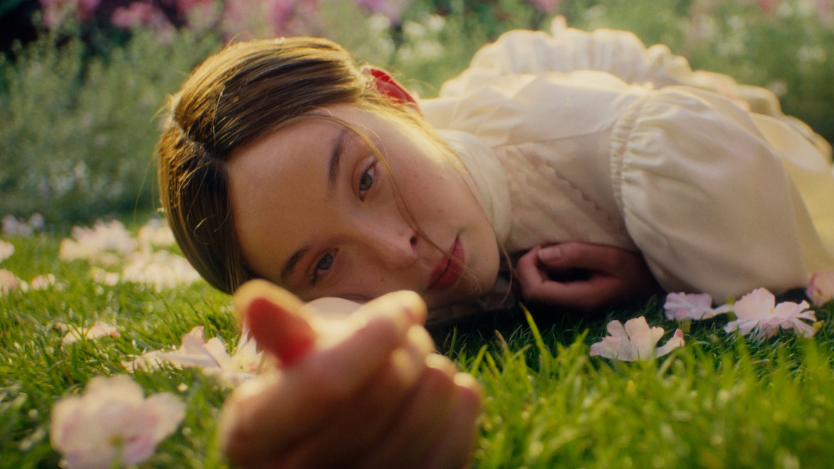A young woman in a light blouse lies on green grass surrounded by pink flowers, looking directly at the camera in a sunlit garden or field.