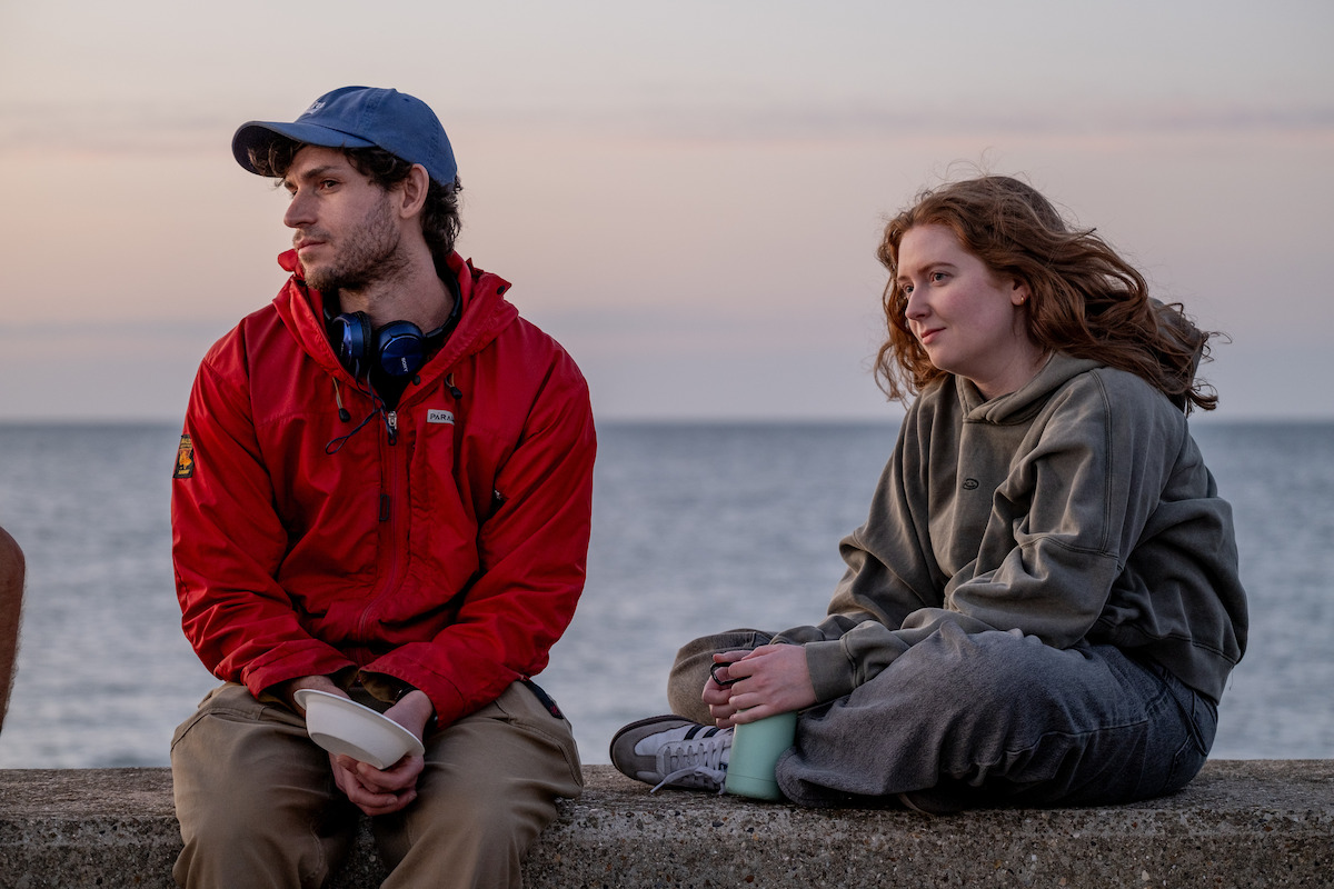 Two people sitting on a stone wall by the sea during sunset, one wearing a red jacket and blue cap, the other in gray casual clothes, both appearing relaxed and having a casual conversation outdoors.