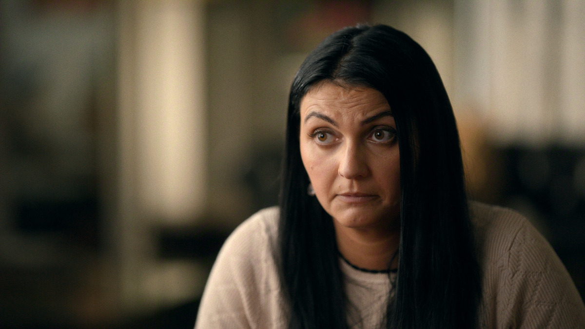 Woman with long dark hair in a light sweater sits indoors, looking slightly skeptical or concerned, with a softly blurred background suggesting an interview or candid conversation in a casual setting.