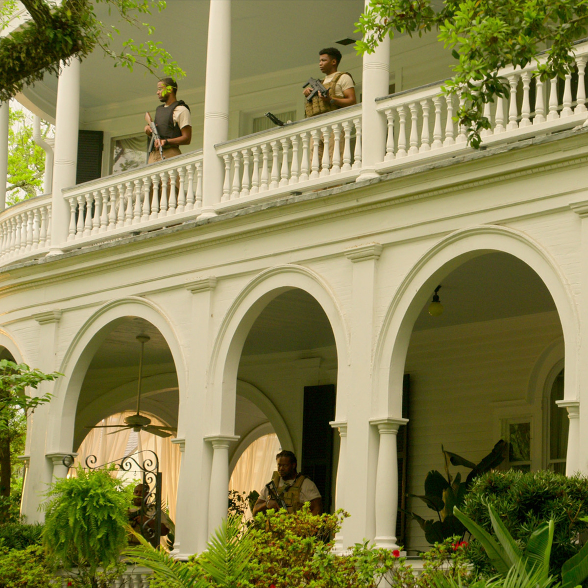 Três pessoas com roupas militares estão em pé na varanda de uma grande casa branca com arcos e colunas, cercados por uma paisagem verde exuberante em um ambiente ao ar livre à luz do dia.