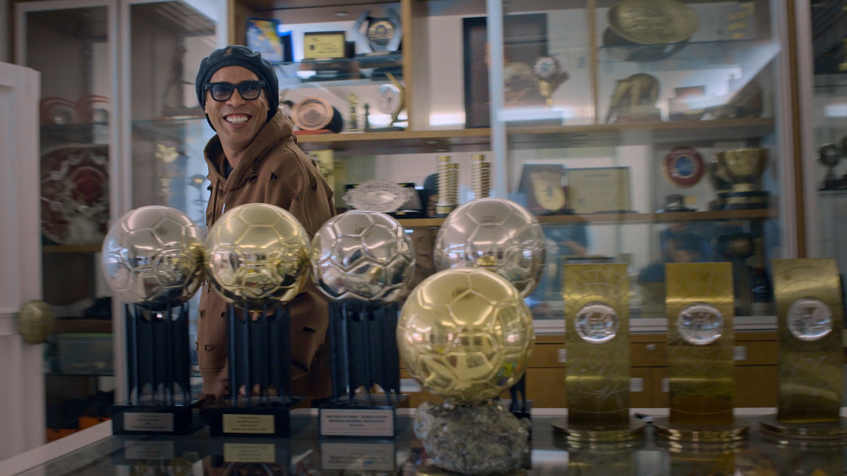 Ronaldinho Gaúcho smiling and wearing glasses stands in a trophy room filled with numerous awards and medals displayed in glass cases and on shelves, with several golden soccer trophies prominently on a table in the foreground.