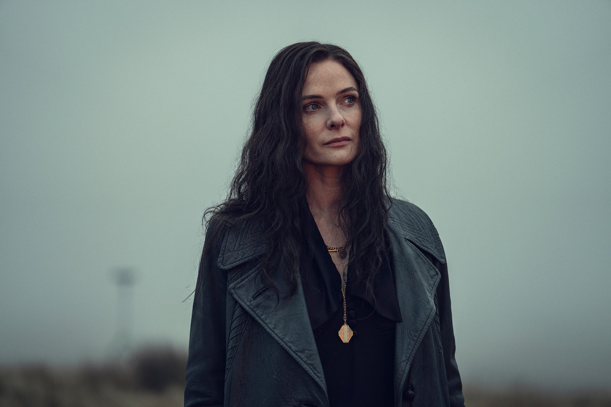 Woman with long dark hair in a black coat standing outdoors on a foggy, overcast day, with blurred grassy landscape and lone structure in the background.