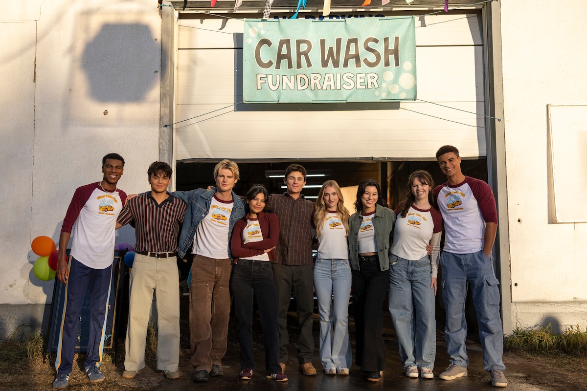 A group of teens stands together smiling outside a garage decorated with balloons and a "Car Wash Fundraiser" sign, posing for a photo in casual clothes, some wearing matching event shirts, during daytime.