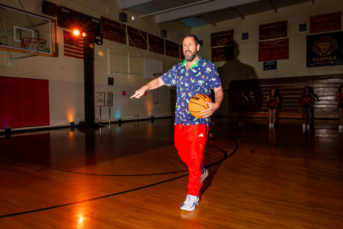 Two people have a conversation on stage in a brightly lit gymnasium, seated in armchairs on a red carpet, surrounded by studio lights, monitors, and a basketball, creating a professional yet relaxed atmosphere.