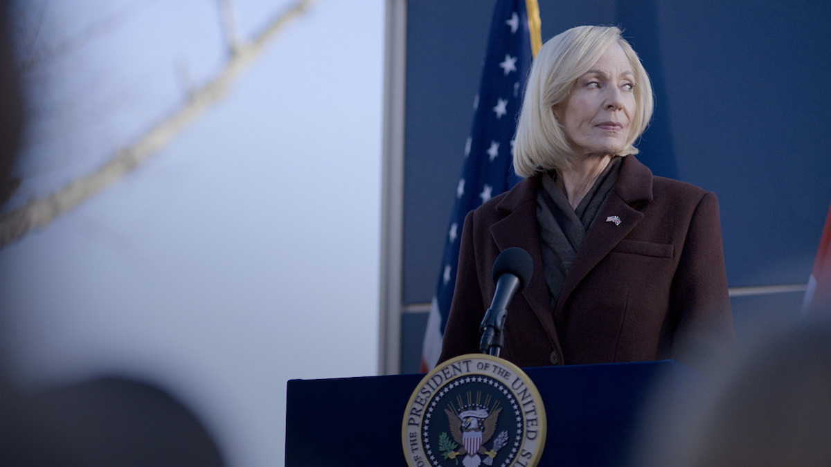 A woman with blonde hair stands at a podium with the seal of the President of the United States, looking to the side, with an American flag behind her.