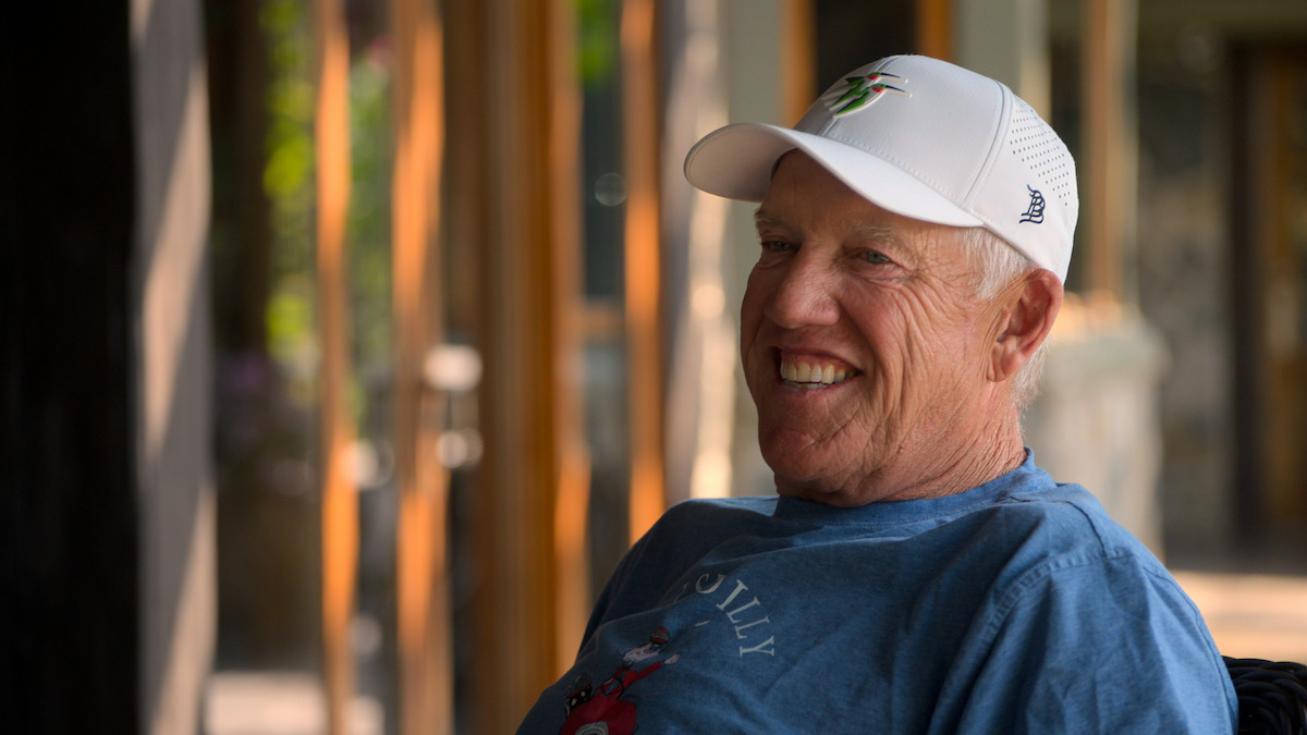 John Elway in a white cap and blue shirt sits outdoors in a bright, shaded area with wooden beams and glass doors in the background, enjoying a relaxed atmosphere.