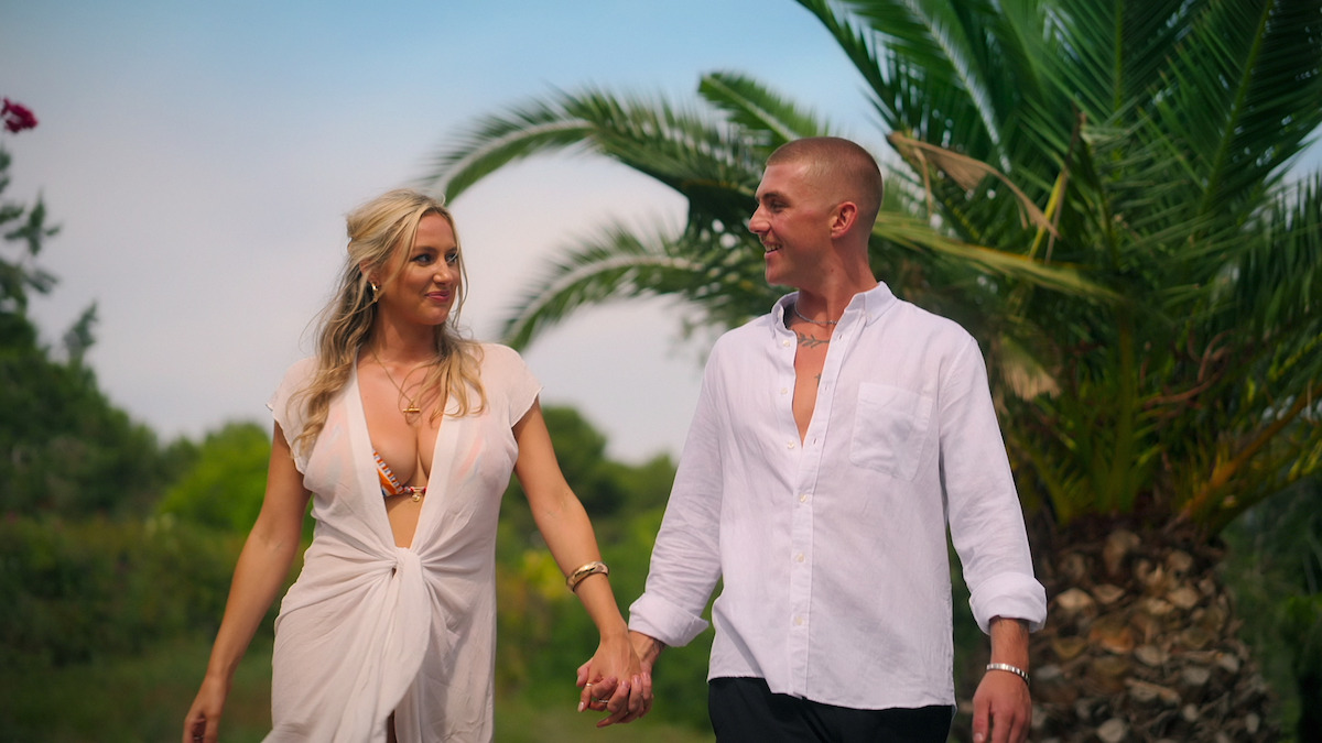 A smiling couple holding hands, walking outdoors in a tropical garden setting with palm trees and greenery, both dressed in light summer clothing, under a partly cloudy sky.