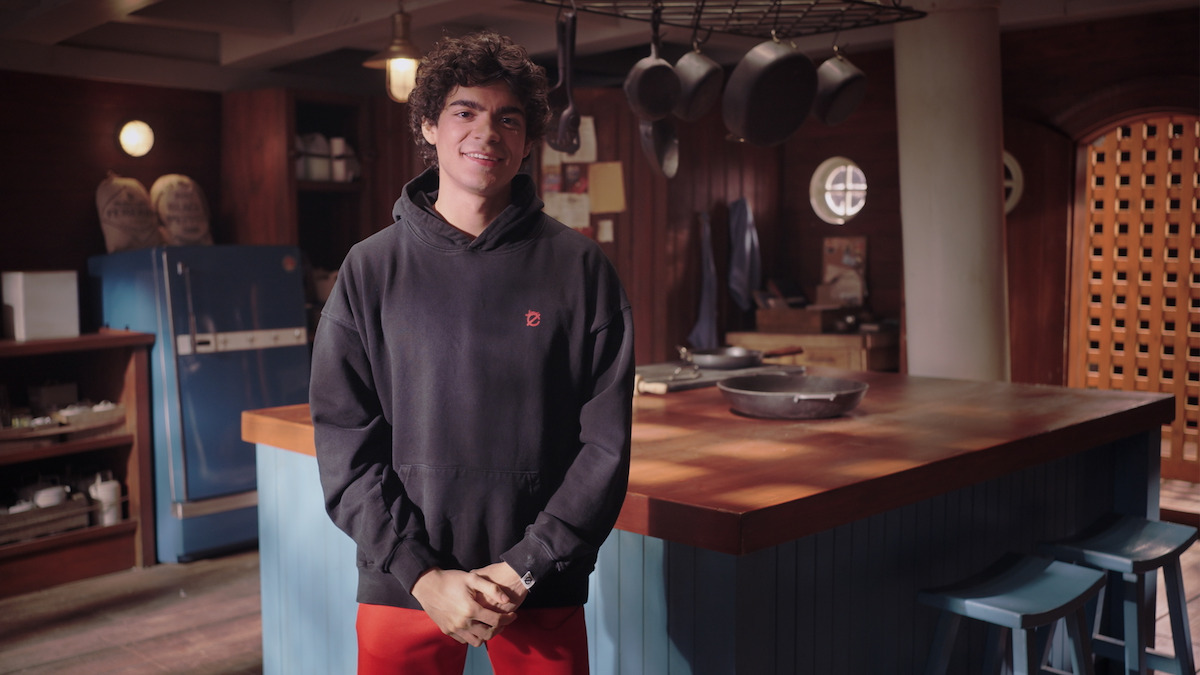 Iñaki Godoy (Monkey D. Luffy in One Piece) wearing a black hoodie and red pants, stands in a cozy kitchen with wooden cabinets, a blue refrigerator, and pots hanging from the ceiling.