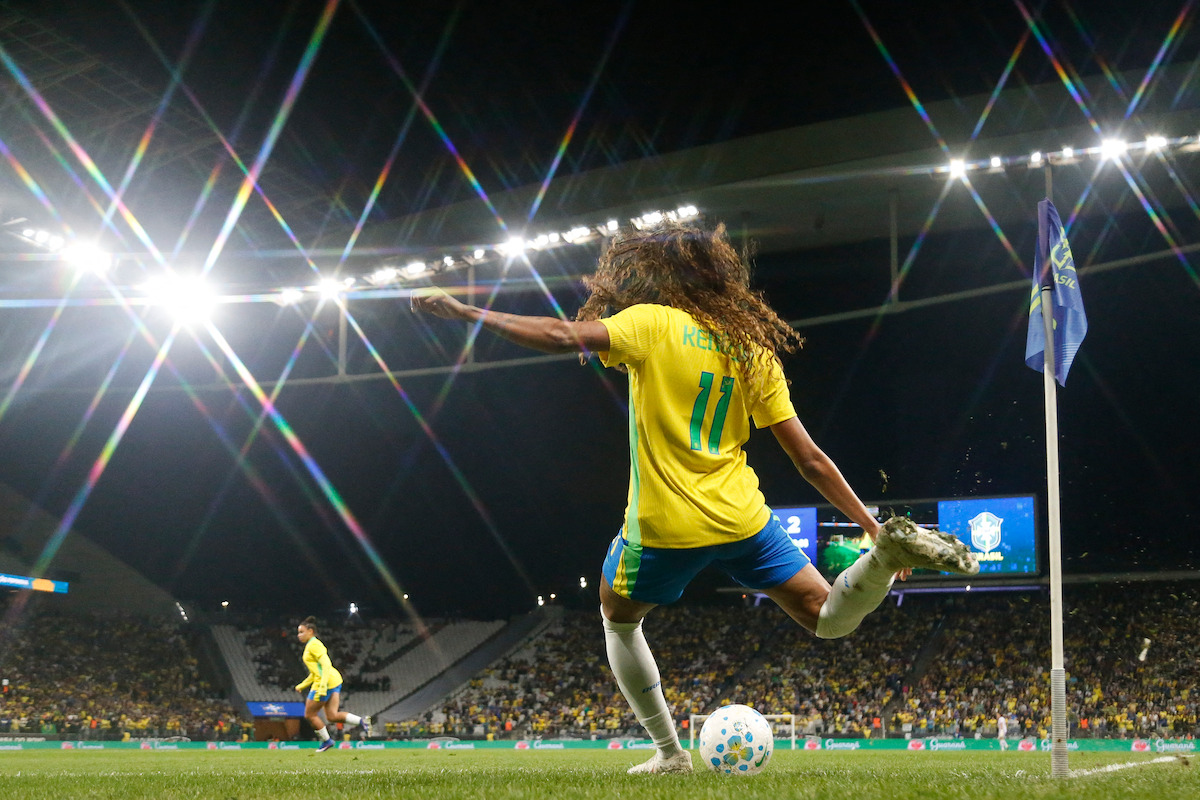 Soccer player in yellow jersey taking a corner kick at night in a stadium, with bright lights, a crowd of spectators, and other players visible in the background.
