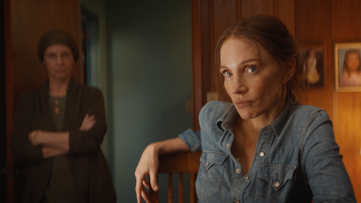 Woman in a denim shirt sits at a wooden table in a warmly lit room, looking serious. Another woman stands in the background with arms crossed. Wooden panel walls and family photos create a cozy, tense atmosphere.
