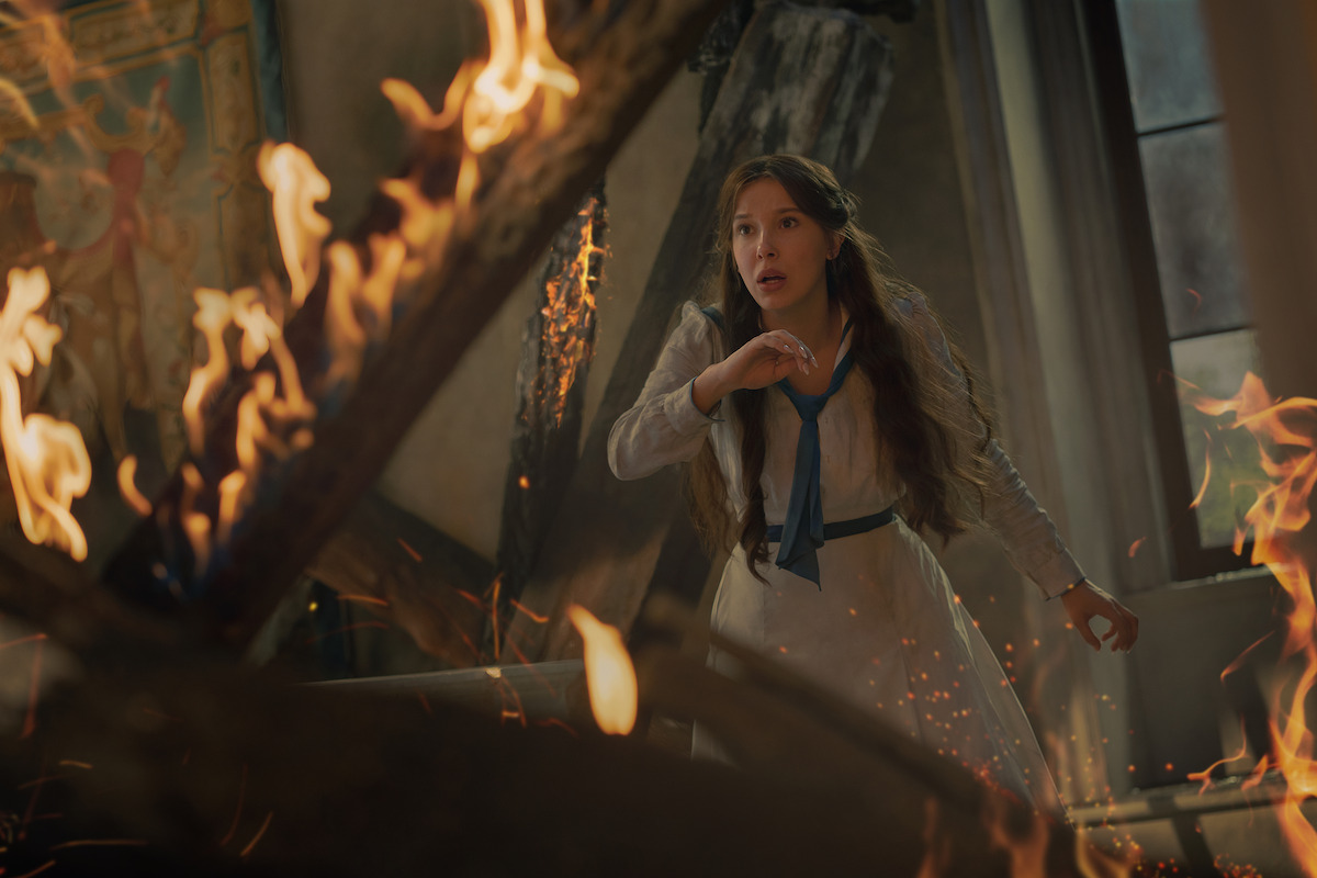Young woman in a white dress stands in a burning room with flames and smoke, looking alarmed and ready to escape, historic or fantasy setting with damaged wooden beams and painted wall in the background.