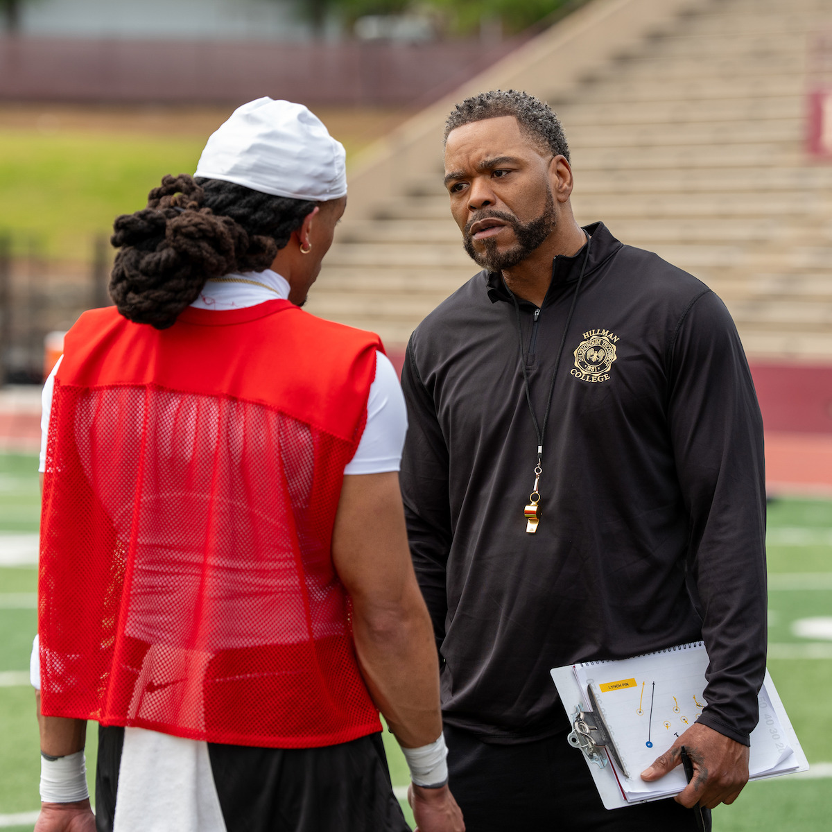Football coach talking to a player on a field, holding a clipboard and whistle, with stadium bleachers in the background, suggesting a practice or training session.