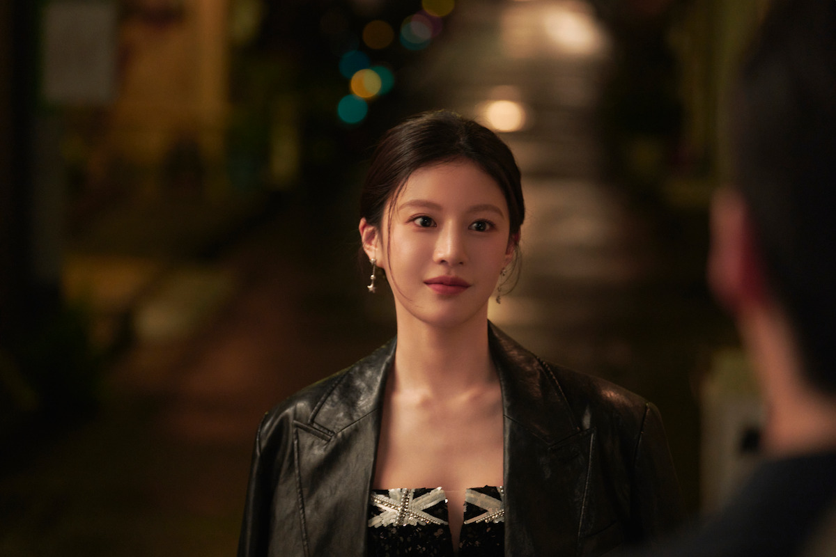 A woman in a black leather jacket stands outside at night on a dimly lit street, looking at someone out of frame with a soft, expectant expression. The mood is intimate and calm, with blurred city lights in the background.