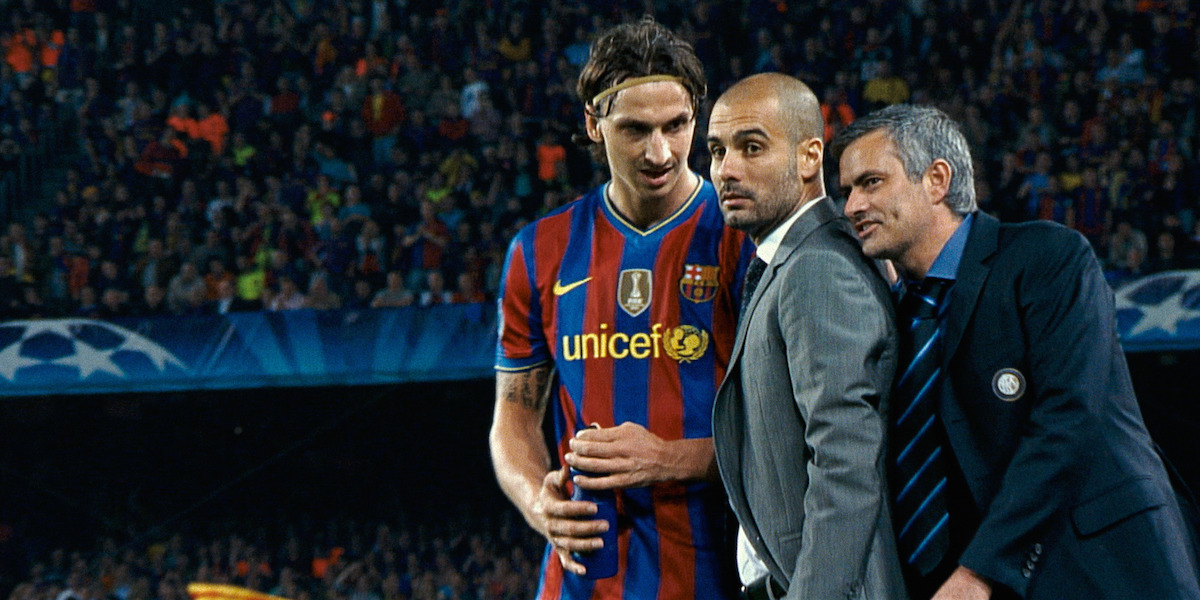 Soccer player in a red and blue jersey with two suited coaches on a crowded stadium field during a high-profile match, appearing to discuss strategy.
