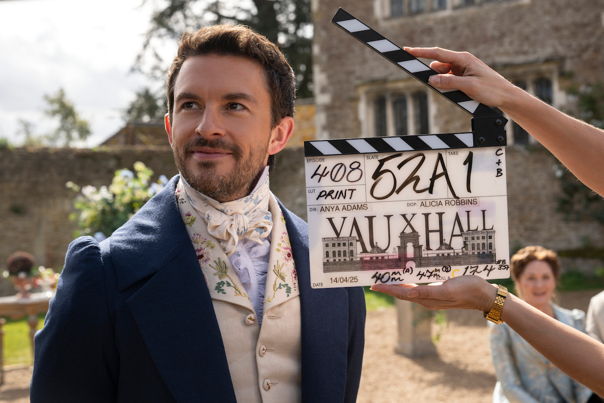 Actor in period costume stands outdoors on set, with a clapboard in front of him. Historic building and people in background, suggesting a film or TV production in a historical setting.