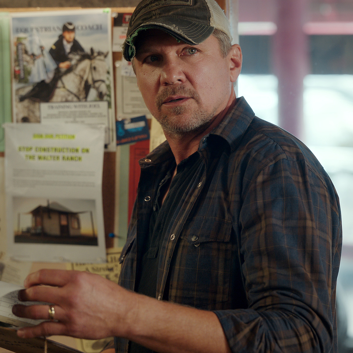 Man in a plaid shirt and cap standing indoors by a bulletin board with papers and flyers, looking surprised or concerned, natural window light in background.