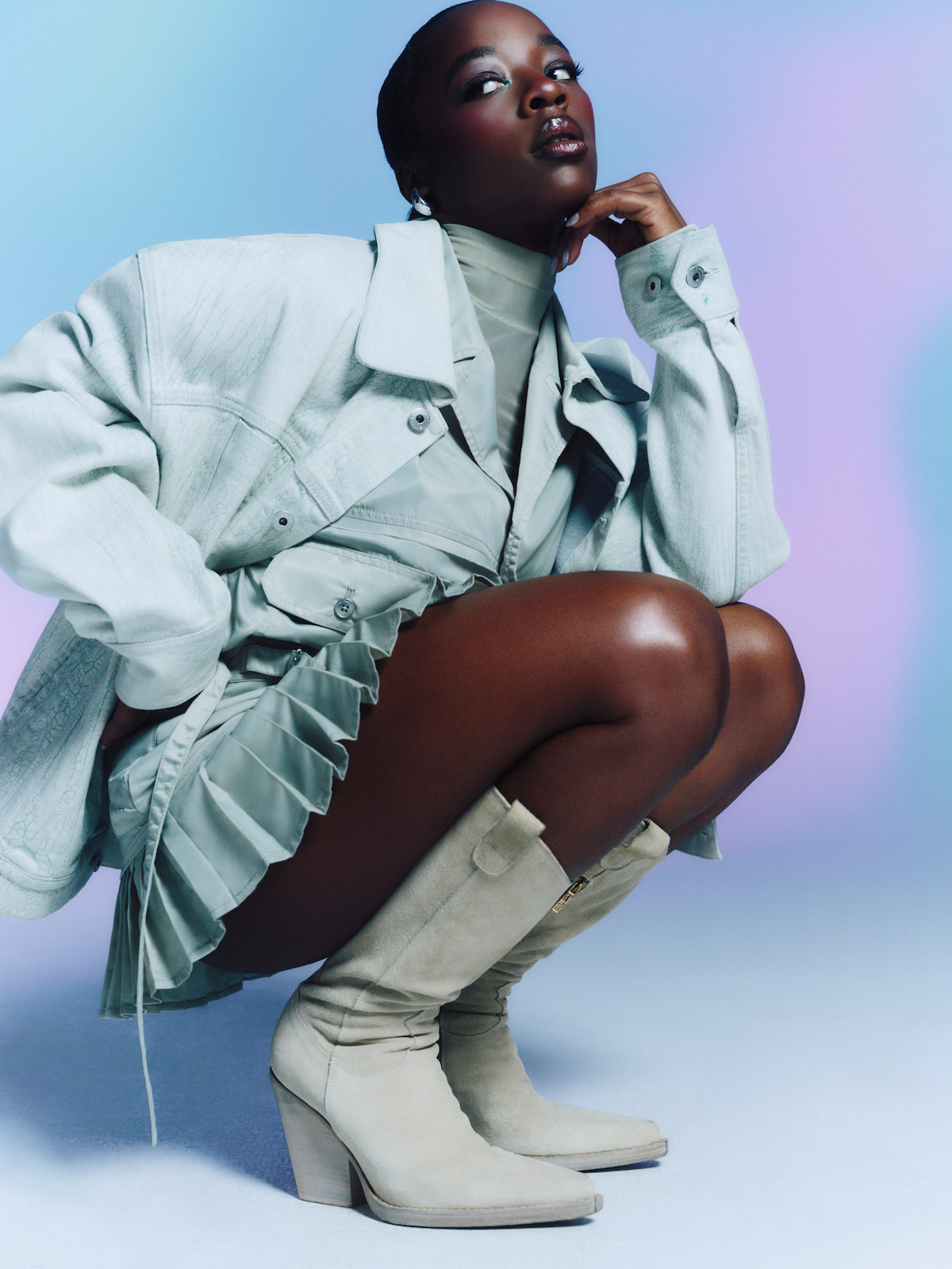 Fashionable woman in light gray jacket, pleated dress, and suede boots poses squatting on one knee, with a thoughtful expression, in a studio with pastel blue and purple gradient background.