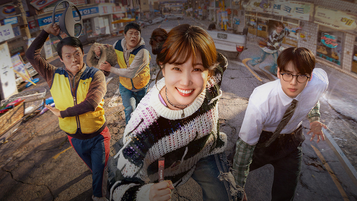 Five people, including a smiling woman in a colorful sweater and a man in glasses, run on a damaged street in a city neighborhood with cracked pavement and rundown storefronts, suggesting urgency or disaster in an urban setting.