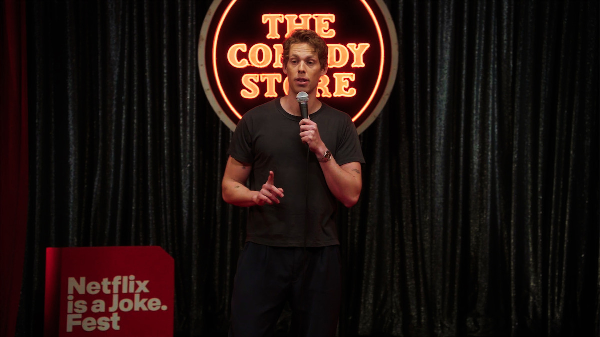 Comedian performs stand-up on stage at The Comedy Store, holding a microphone. Behind is a neon sign and dark curtains. A red podium to the left displays the text "Netflix is a Joke Fest."