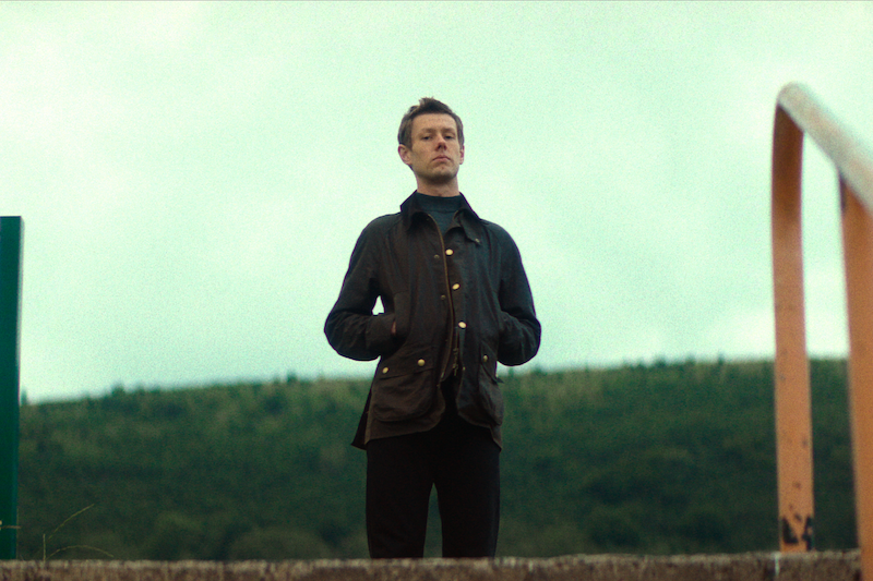 A man stands outdoors with hands in coat pockets, looking serious. He is on a concrete platform, flanked by a metal railing, with a backdrop of green hills under a cloudy sky. The scene appears calm and slightly overcast.