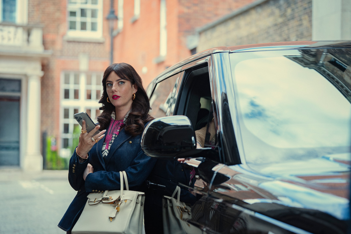 Woman with long dark hair in a stylish blazer stands next to a black luxury car on a city street, holding a smartphone and beige purse, with upscale buildings in the background.