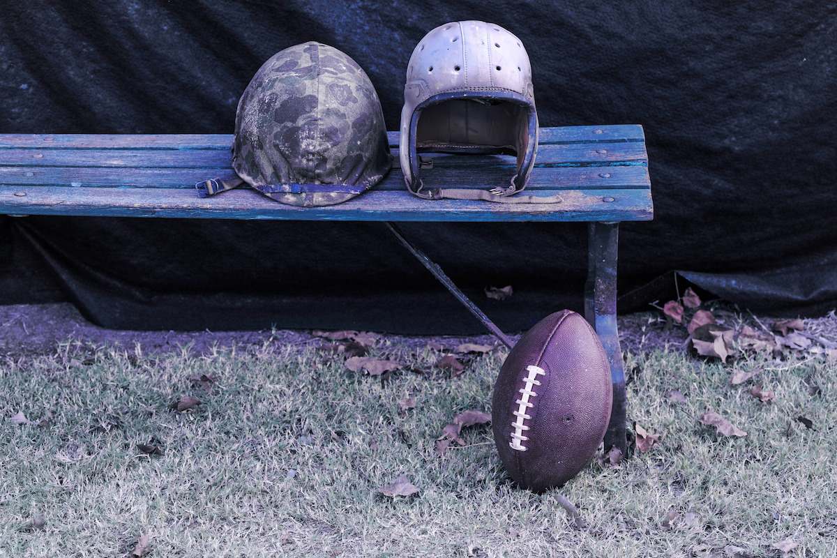 Behind-the-scenes photo of an army helmet and a football helmet on a bench on the set of ‘The Mosquito Bowl.’