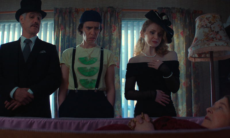 Three people stand in a dimly lit room by a window, dressed in funeral attire, looking solemnly at a woman lying in an open casket, creating a somber mood.