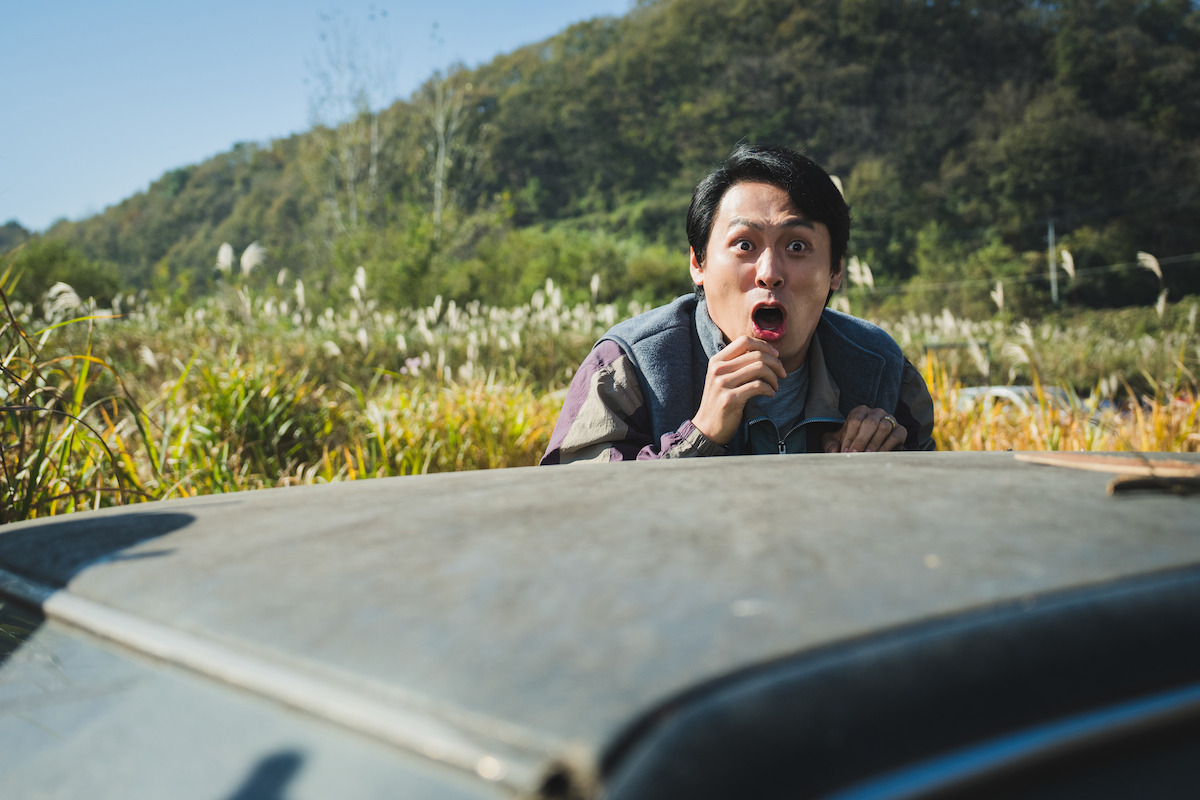 A surprised man standing behind a car in an outdoor rural setting with tall grass and trees in the background, under a clear blue sky.
