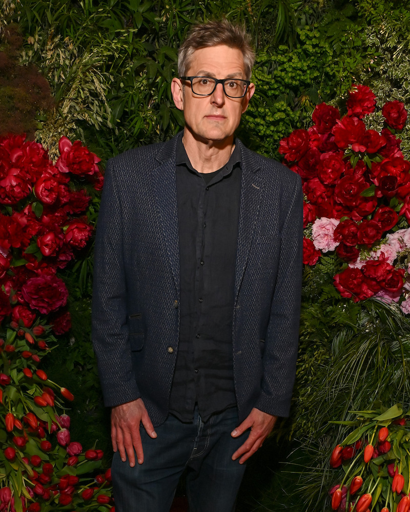 Man with glasses wearing a dark jacket and shirt stands in front of a vibrant floral backdrop filled with red flowers and greenery. Indoor setting with dramatic, colorful decorations.