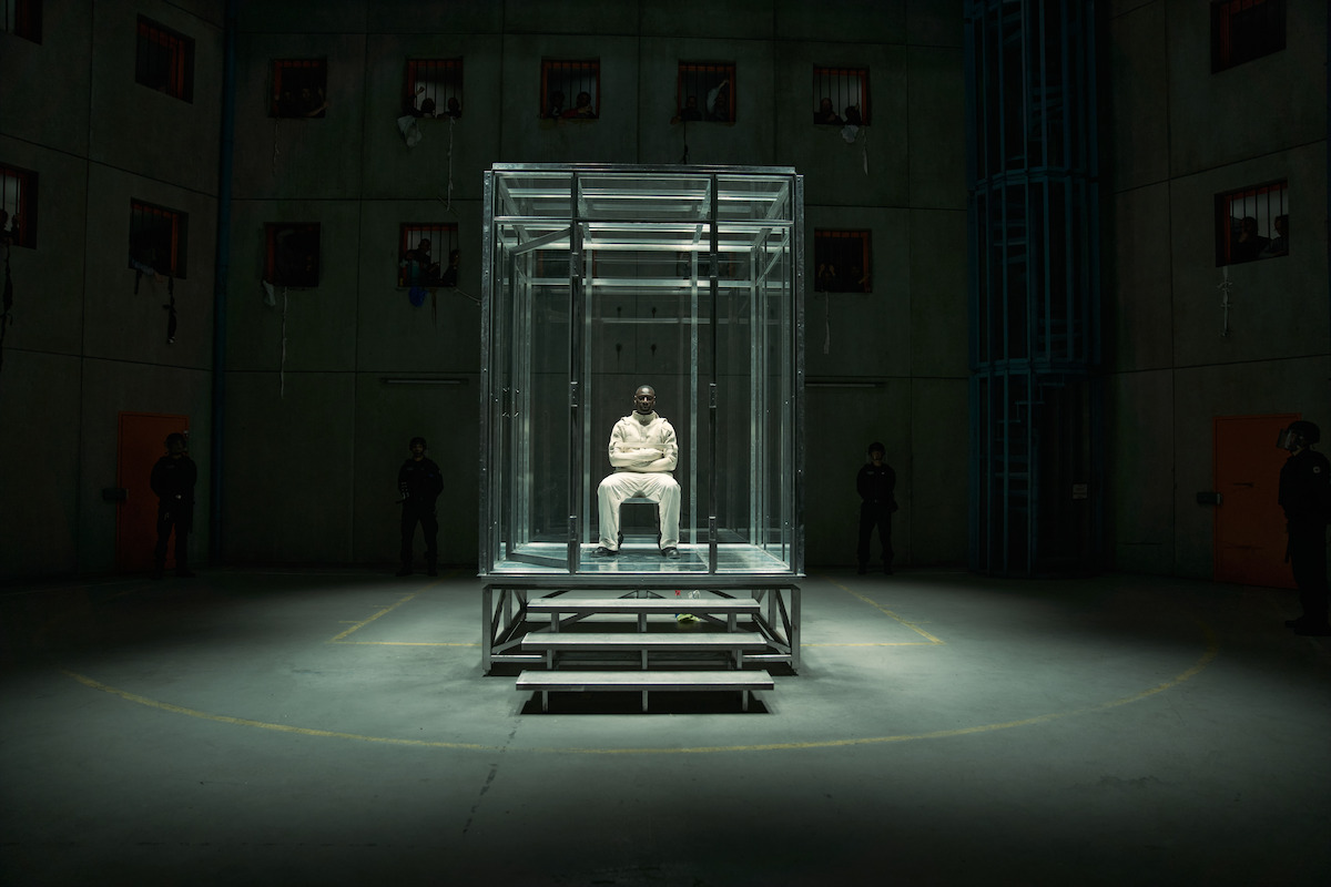 A person in a white outfit sits alone inside a glass cell at the center of a dimly lit, industrial room with concrete walls. Several shadowy figures stand in the background, emphasizing a tense, isolated prison environment.