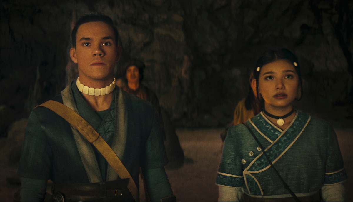 Two young people in blue traditional clothing stand in a dimly lit cave, looking ahead with serious expressions. Stone walls and another figure are visible in the background, suggesting a tense or significant moment.