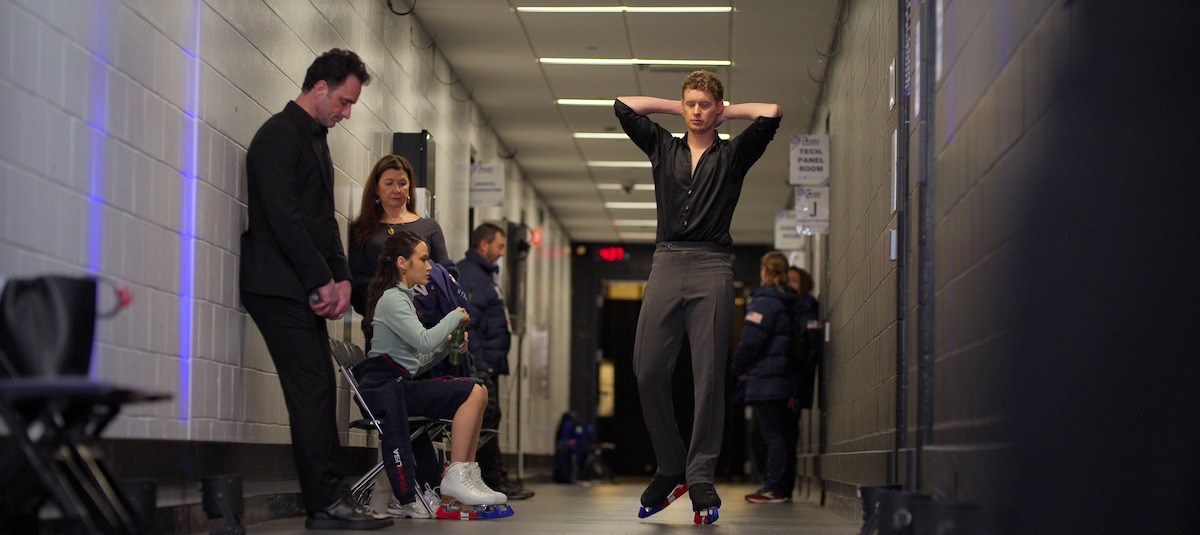 Patrice Lauzon, Madison Chock, and Evan Bates wait and prepare in a brightly lit hallway backstage at an ice rink, with benches, gear, and skates visible along the corridor.