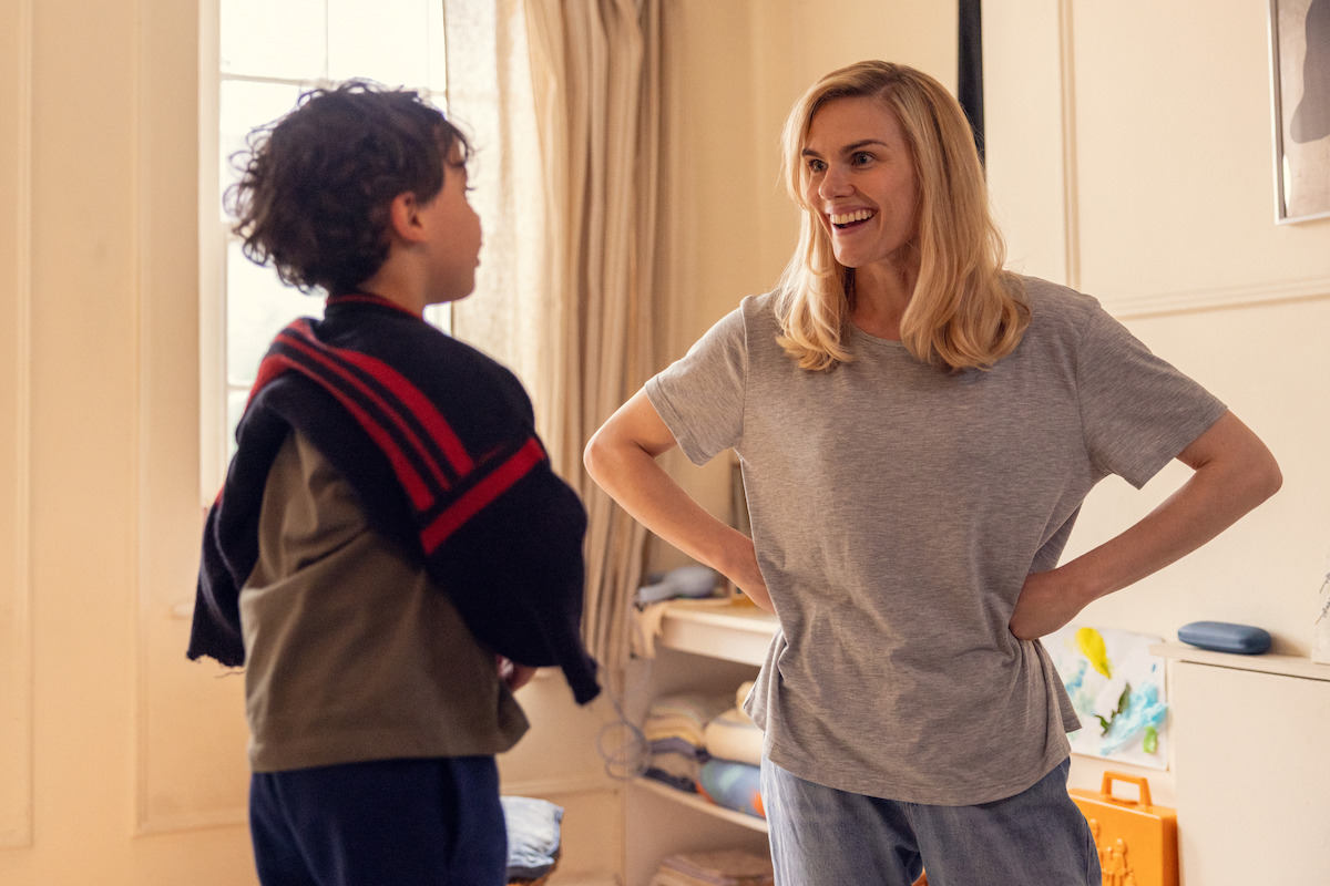 A smiling woman talks to a young boy in a cozy, sunlit room with shelves, toys, and a window with curtains, creating a warm, homey environment.