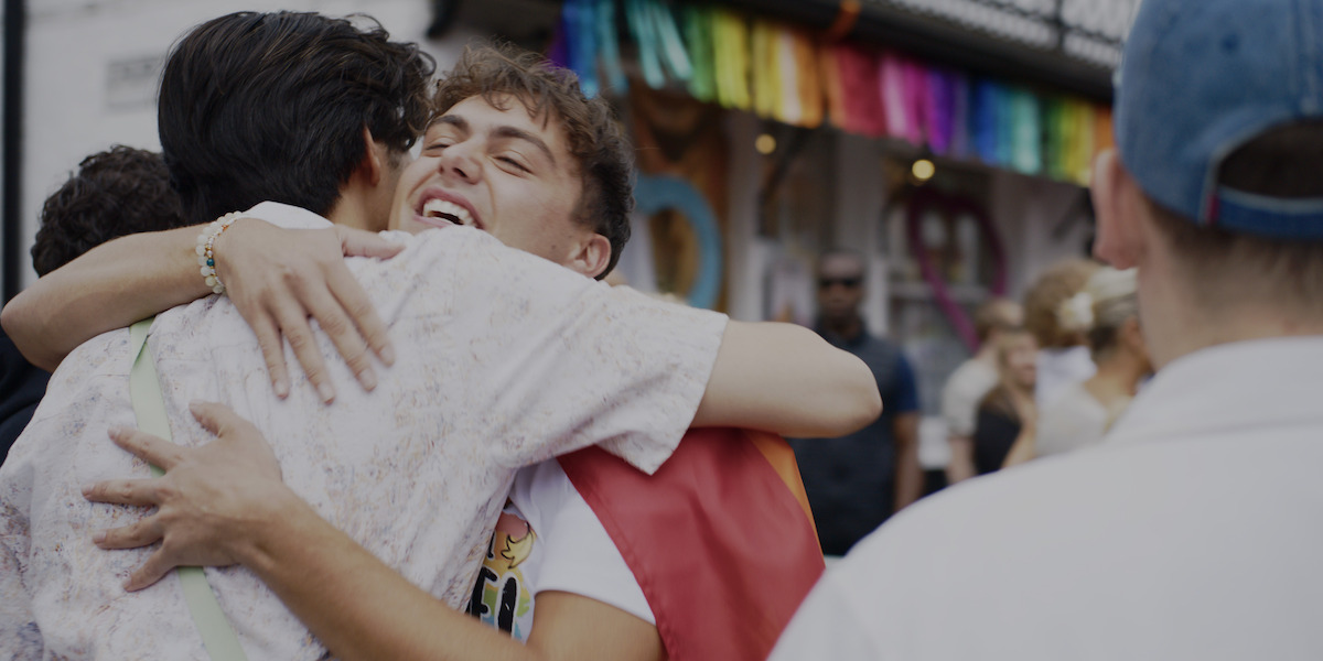 Two people hugging and smiling at an outdoor event with a rainbow banner in the background, suggesting a celebration or pride festival, surrounded by other attendees.