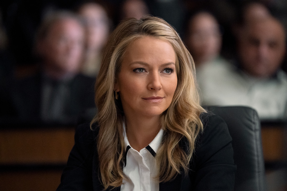 A woman with blonde hair wearing a black blazer and white shirt is seated at a table in a formal setting, likely a courtroom or meeting room, with blurred people in the background watching attentively.
