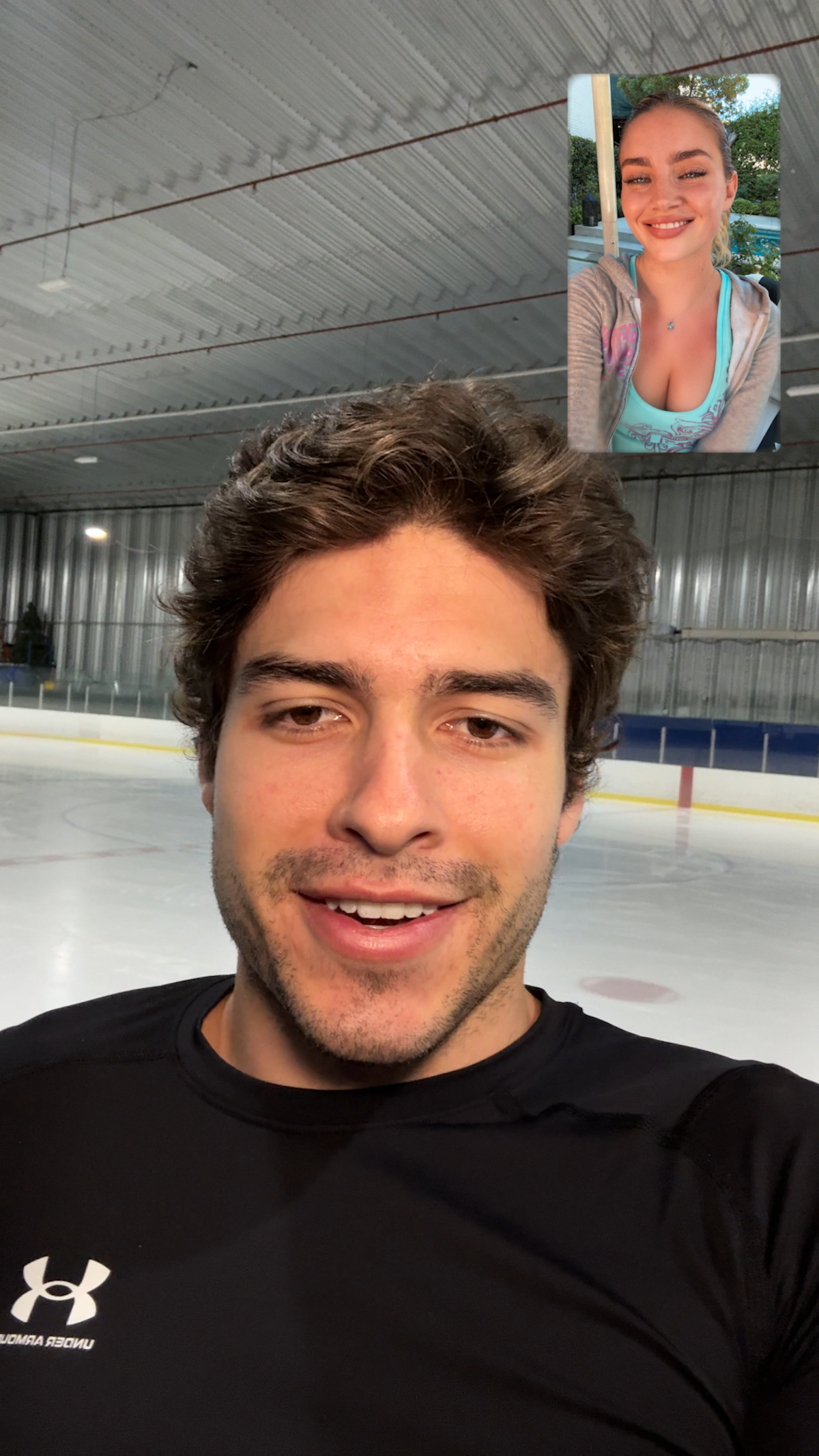 Man wearing a black sports shirt taking a selfie video call at an indoor ice rink, with a smiling woman appearing on the screen in the top corner, possibly outdoors.