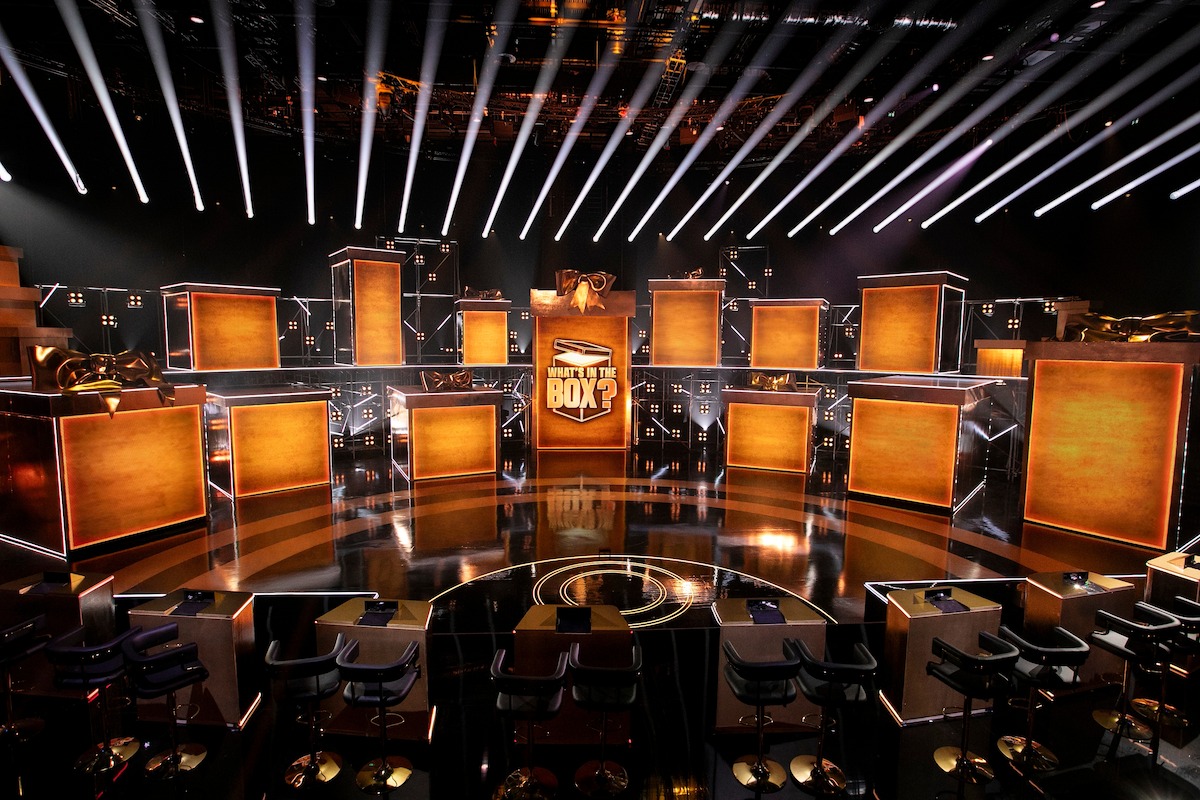 A dramatic television game show set with illuminated orange boxes, a central stage with a logo, dramatic spotlights above, and audience seating, creating an intense and energetic studio atmosphere.