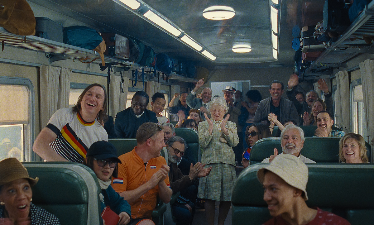 A diverse group of smiling and cheering passengers sits and stands inside a brightly lit, vintage train car, creating a joyful, celebratory atmosphere.