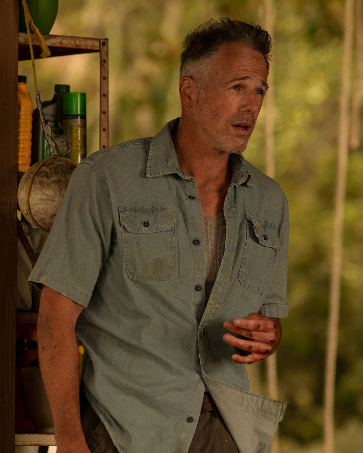 Gary Weeks as Luke Maybank with short hair in a light denim shirt stands near shelves outdoors, looking concerned; his hand is stained with blood, with various tools and bottles behind him and greenery in the background.