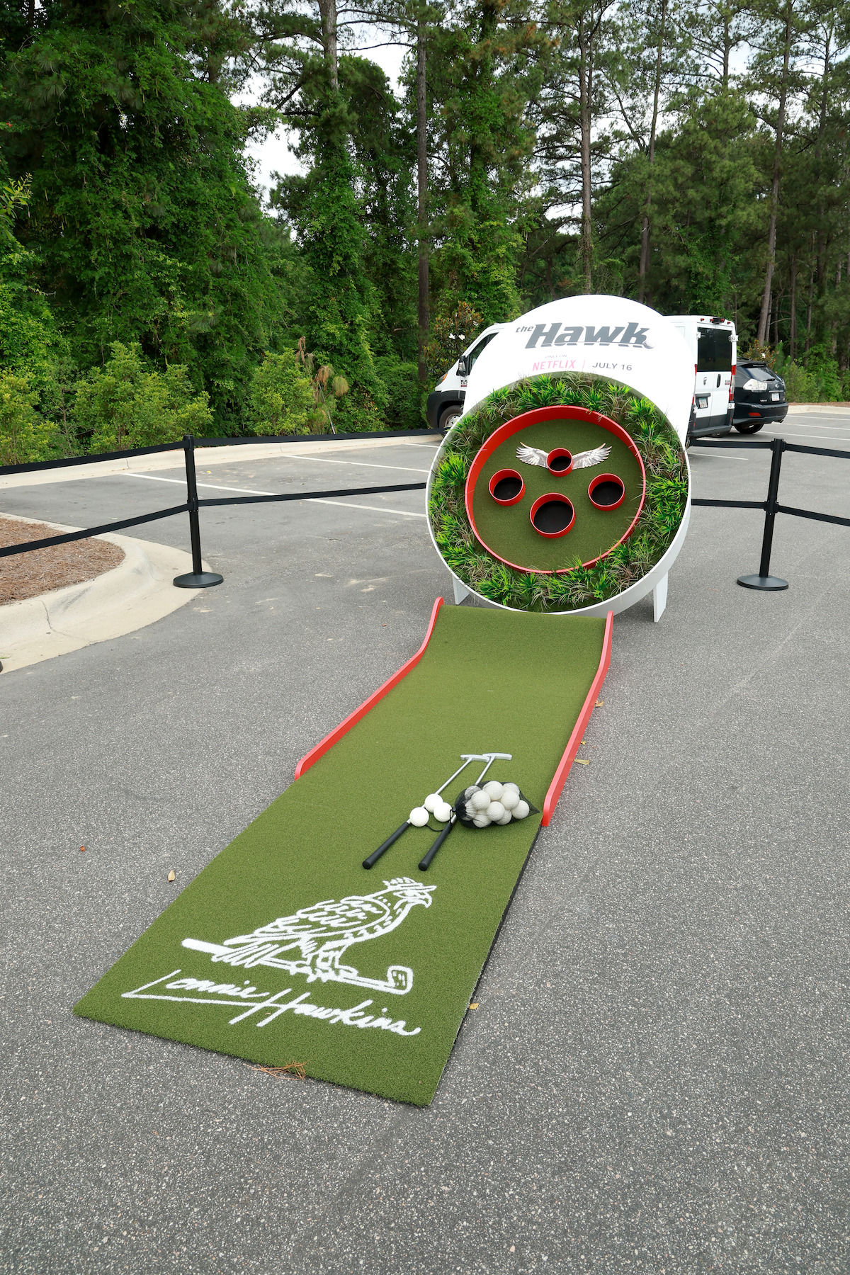 Mini-golf putting green with golf clubs and balls, leading to a large circular target with holes, set up outdoors on a street near greenery and parked vehicles, surrounded by black ropes for an event or activity.