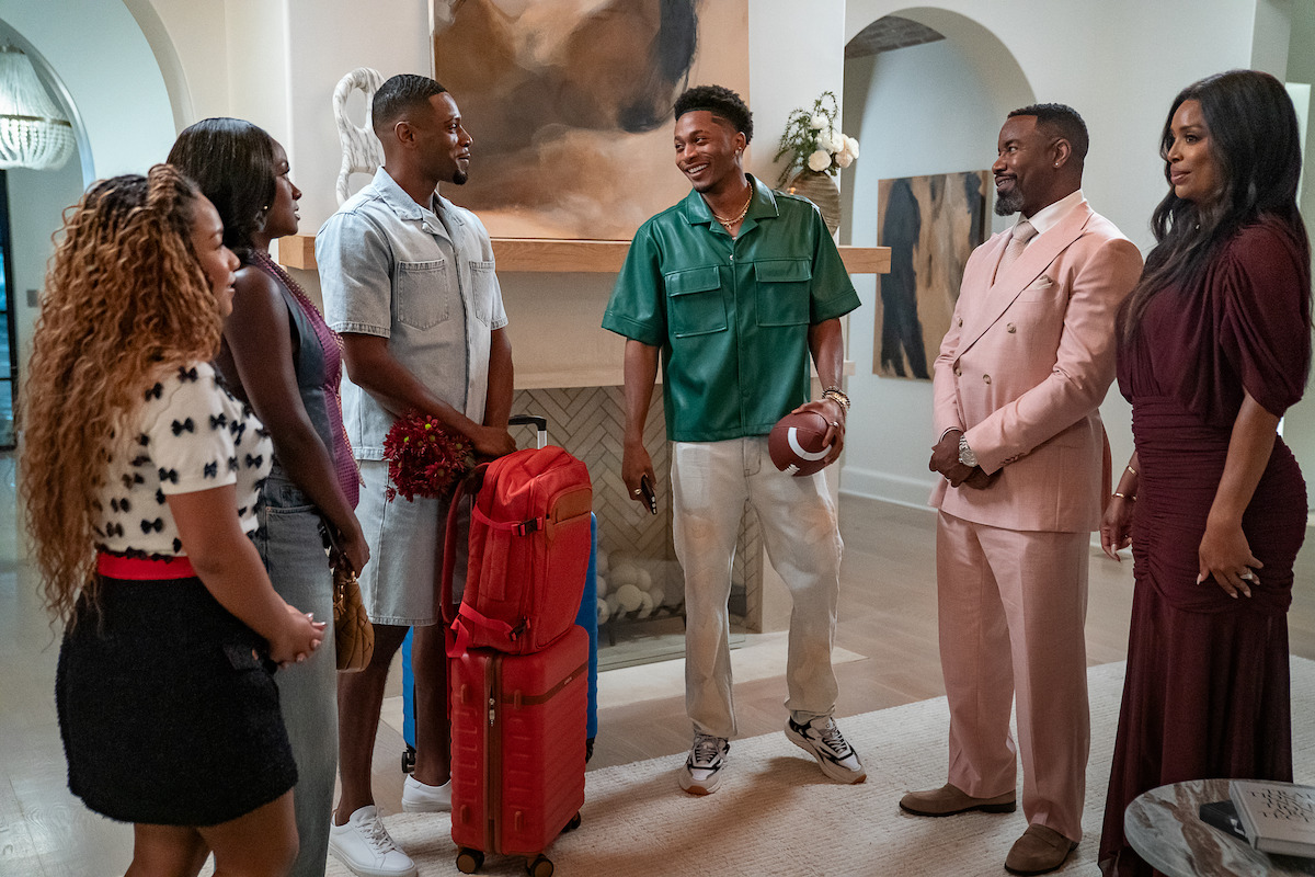 Six people gather in a stylish, modern living room. One person holds a football and luggage, suggesting arrival or departure, while others smile, dressed in a mix of casual and formal outfits. Bright, elegant home interior.