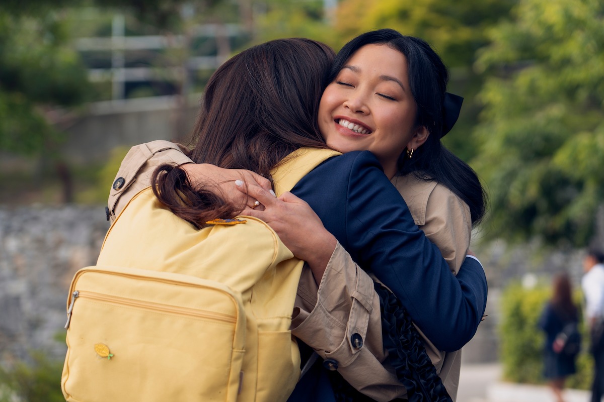 Two young women outdoors hugging warmly, one wearing a yellow backpack, both smiling in a green park setting with blurred people in the background.