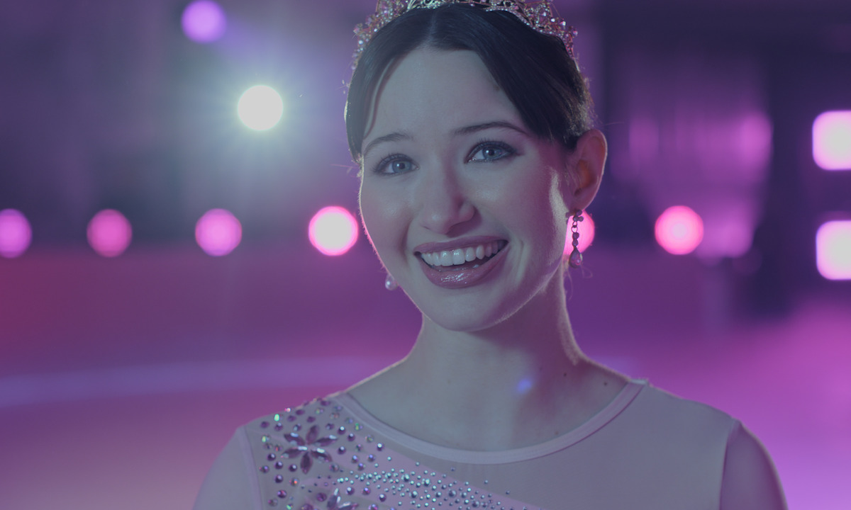Smiling young woman with a tiara and jeweled dress, standing under purple stage lights in an indoor setting, possibly at a performance or celebration event.