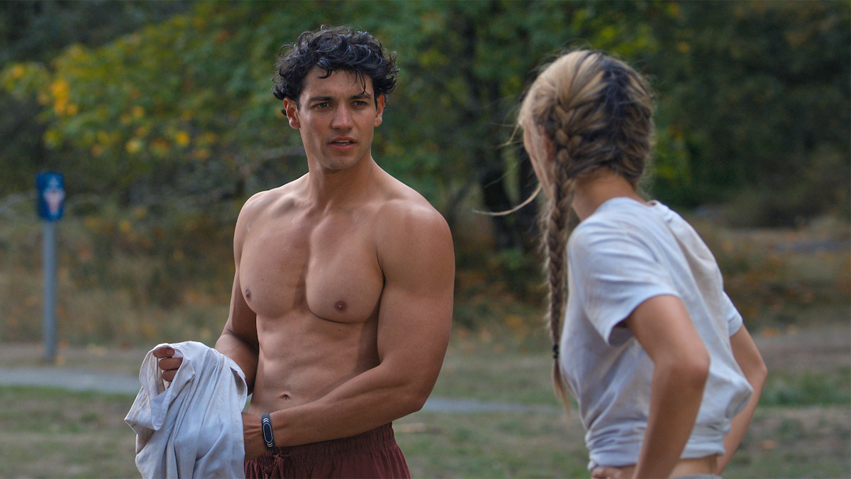 A shirtless man holding a shirt talks to a woman with braided hair in a casual outdoor setting with grass and trees, suggesting a relaxed or recreational environment.