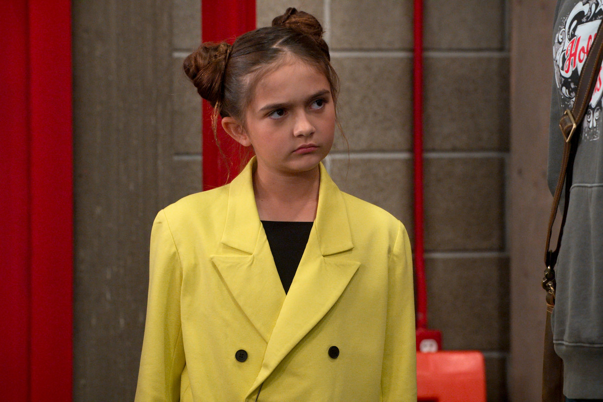 Young girl with brown hair in two buns, wearing a bright yellow jacket, stands indoors against a wall with red accents and looks to the side with a serious expression.